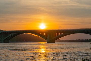 Fototapeta premium A captivating image featuring a suspension bridge over water, showcasing architectural elegance and natural beauty as the sun sets - Kyiv, Ukraine 06-17-2024
