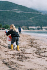 Little girl puts a bottle in a bag of trash on a sandy beach. Back view