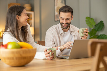 A couple enjoys drinks together, laughing and interacting over a laptop in a cozy kitchen, showcasing love and happiness.