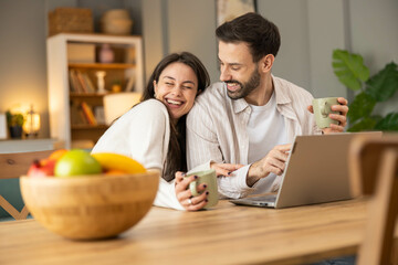 A couple sits together sharing smiles, drinks, and laughter while engaged with a laptop in a cozy kitchen environment.