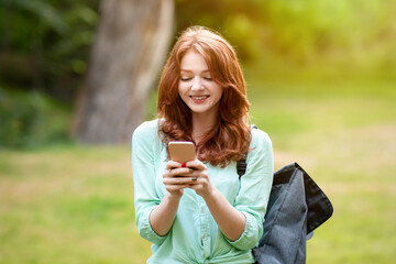 Fototapeta premium Cheerful redhead student girl using smartphone outdoors, texting with friends, standing at college campus yard with backpack