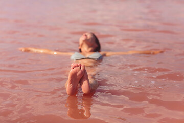 A mature woman floats on her back in a pink saltwater lake, with the camera sharply focused on her feet above the water, creating a unique perspective under a clear sunny sky.