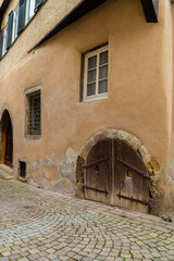 Fototapeta premium Old building facade with a rounded wooden door and two windows. Cobblestone street, weathered stucco wall. European architecture, historic detail.
