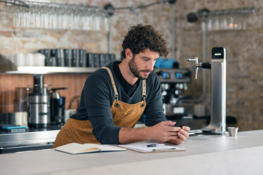 Handsome waiter taking inventory of everything missing while making a checklist on his phone at the coffee shop - Powered by Adobe