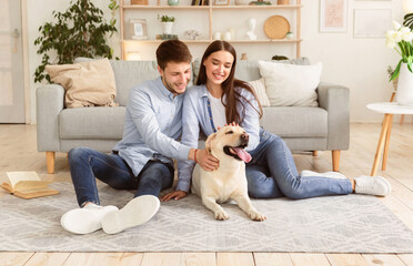 Happy Loving Family. Portrait of beautiful spouses patting dog sitting on the floor carpet in...