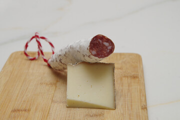 a wedge of cured sheep's cheese with a moldy rind and a piece of salami with its cord on a wooden cutting board on a white-veined marble table