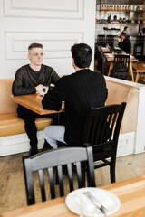 Fototapeta premium A vertical medium shot of a gay men couple on a date in a cozy cafe A mid shot of a LGBTQ+ couple on a romantic date, sitting by a table and talking in an urban cafe interior