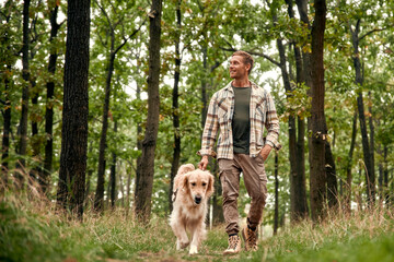 A man is leisurely walking his cheerful Golden Retriever through a lush, beautiful forest area