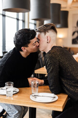 A vertical medium shot of a gay men couple on a date in a cozy cafe
A mid shot of a LGBTQ+ couple on a romantic date, a man timidly touching his partner's hand and kissing him across a table in an urb
