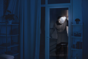 Criminal Act Concept. Masked robber looking at apartment through balcony door, using flashlight, copy space