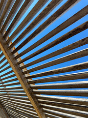 Low angle view of a wooden pergola with parallel slats creating a geometric pattern against a clear blue sky.
