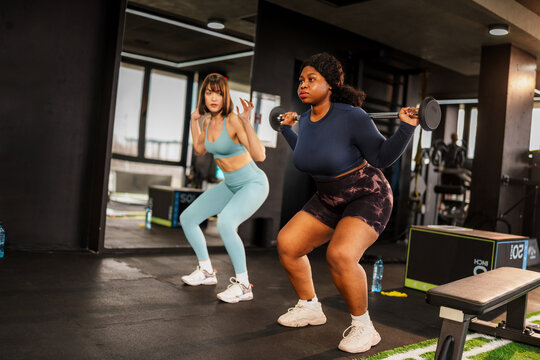 Two female athletes performing squats with weights in a brightly lit gym, showcasing strength and dedication to their fitness journey while embodying a powerful and active lifestyle