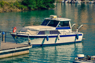 A white and blue motorboat is moored to a wooden pier in a calm harbor on a sunny day, evoking a...