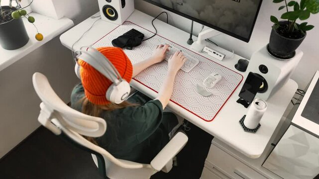 Comfortable workspace with modern gaming setup. Girl in beanie and headphones types on white keyboard while focusing on screen. Stylish and organized desk setup enhances efficiency and concentration.
