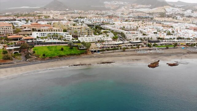 Aerial view of Costa Adeje city, Tenerife, Canary islands. Cinematic 4K footage with sandy coast, calm waters of Atlantic ocean, city modern villas, quay with people, mountains. Early morning video