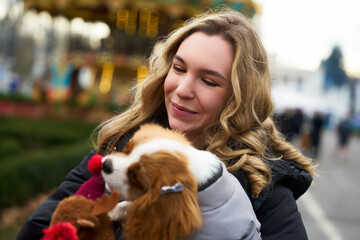 Woman carrying a small dog during a winter festival in a city park with festive attractions and people in the background