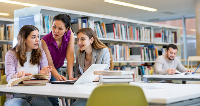Three girls students, preparing for classes on a laptop in the university library, are looking for information in textbooks