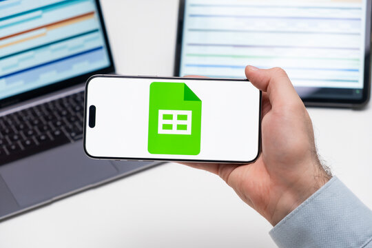 Google sheets logo on the screen of a smartphone in mans hand with laptop and tablet with graphical diagrams on the background, December 2023, Prague, Czech Republic.