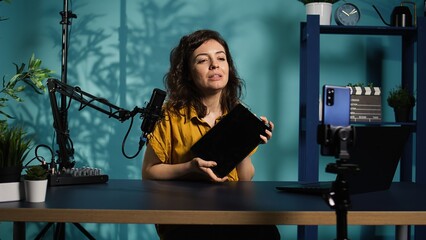 Tech expert presenting tablet in entry level low budget price range, filming from dimly lit home studio. Viral tech content creator reviewing digital device in front of online audience, camera B