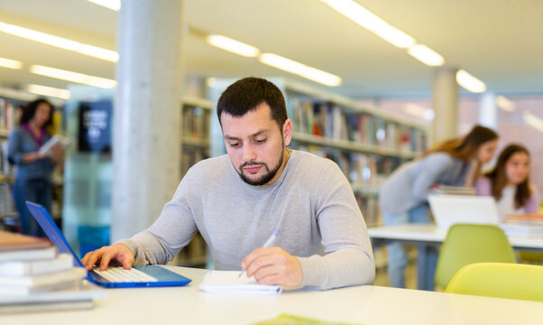 Focused guy student studying at the university studies in the library on a laptop while preparing for the exam, making ..important notes in a copybook