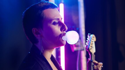 Cool grunge artist blowing bubbles with bubble gum flavor in studio, chewing gum and acting tough. Female musician rocker with leather jacket performs alternative music. Camera A.