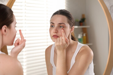 Young woman putting in red color contact lens near mirror indoors