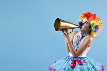 Happy girl dressed like clown with megaphone on light blue background, space for text. Surprise...