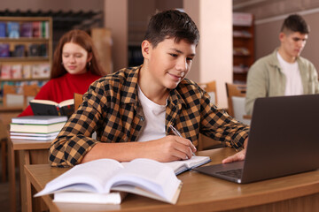 Boy taking notes while using laptop at desk in public library