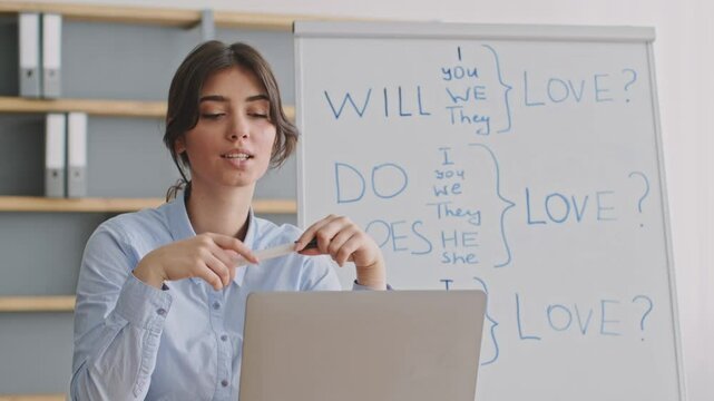A young woman is leading an online educational session, using a laptop and a whiteboard at her home. She explains English verb conjugations, engaging her students remotely.