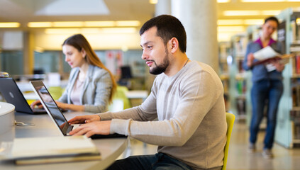 Interested young man working on science project in public library, looking for information in books and websites on laptop..