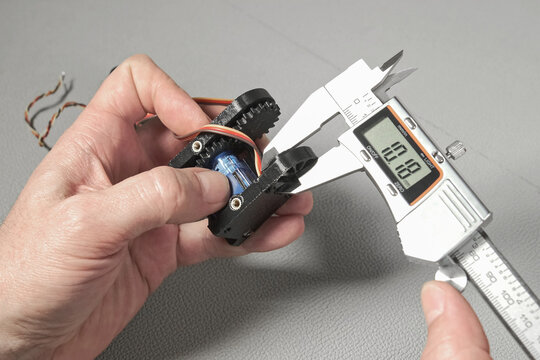 An engineer works with a digital caliper to measure a small robotic component with gears and wiring. The precise measurement is shown on the LCD screen of the tool.