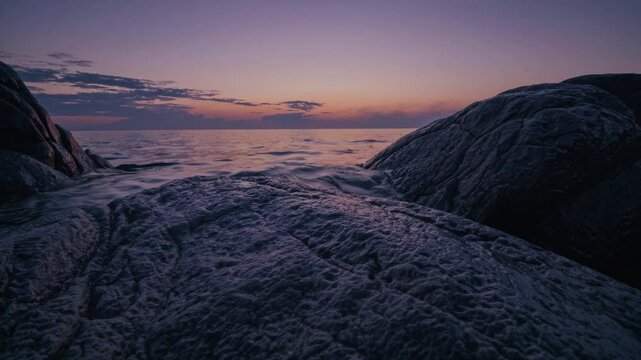 A Serene Coastal Sunset Scene Featuring Smooth, Dark Rocks in the Foreground &ndash; Calm Waters Stretching Toward the Horizon, with Warm Golden and Pink Hues Painting the Sky, Capturing the Tranquility