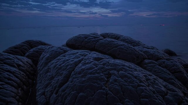 A Serene Coastal Sunset Scene Featuring Smooth, Dark Rocks in the Foreground &ndash; Calm Waters Stretching Toward the Horizon, with Warm Golden and Pink Hues Painting the Sky, Capturing the Tranquility 