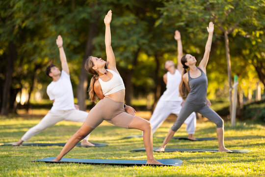 People group in park practicing yoga outdoor. Active pastime together, performing gymnastic exercise. Yoga lesson visitors perform warrior pose. People practice sports during yoga in nature