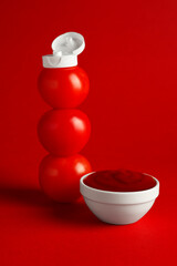 Stack of fresh tomatoes with plastic cap as bottle and ketchup in bowl on red background