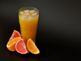 A tall, faceted glass of fruit juice with ice on a black background, next to ripe orange and...