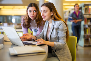 Two young positive women working together on joint project in library, finding information in books and Internet..