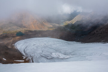 Misty top view from big round glacier to green rocky hilly valley with few turquoise mountain lakes under low clouds. Several most beautiful glacial lakes in low clouds in foggy changeable weather.