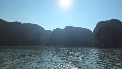 A serene view of Halong Bay in Vietnam, surrounded by towering limestone mountains under a bright sun, with shimmering water reflecting the sunlight.