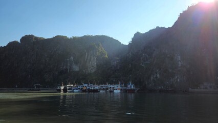 A scenic view of a tourist pier in Ha Long Bay, Vietnam, near Thien Cung Cave, with cruise boats docked by towering limestone mountains under the warm glow of the sun.