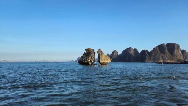 Rocks of the Kissing Cocks in Ha Long Bay, Vietnam, with the limestone formations rising from the sea against a backdrop of majestic karst mountains under a clear blue sky.