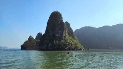 A towering limestone island covered in lush greenery rises from the emerald waters of Ha Long Bay, Vietnam, under a clear blue sky.