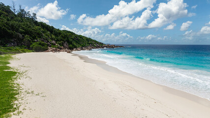 Turquoise ocean waves gently meeting a white sandy beach surrounded by lush green vegetation. Petite Anse. Seychelles, La Digue.
