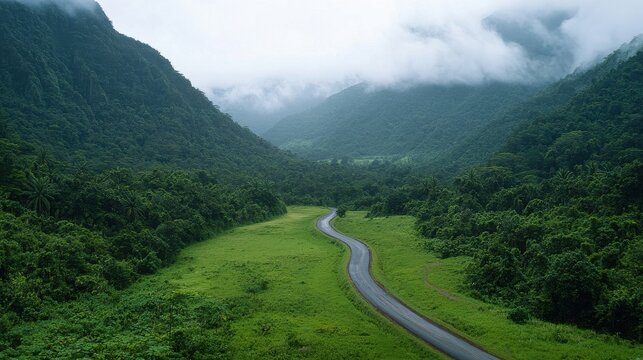 A winding road meanders through lush green valleys, surrounded by towering mountains and misty clouds, creating a serene natural landscape.