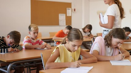 Focused preteen pupils studying in classroom with female teacher, writing exercises in workbooks