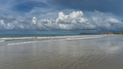 Beautiful tropical beach during the day. The waves of the turquoise ocean are foaming, spreading across the sand. The hotel building, the hills in the distance. Picturesque cumulus clouds, blue sky.  