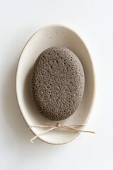 Photo of a round, dark brown pumice stone on its side with a textured surface and small holes throughout, sitting in an oval-shaped, cream-colored dish against a white background. 