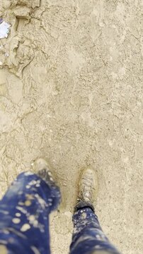 Close-up of legs walking through thick mud after a severe flood. The rich brown tones and squelching steps highlight the raw aftermath, focusing on the texture and movement of the muddy ground.