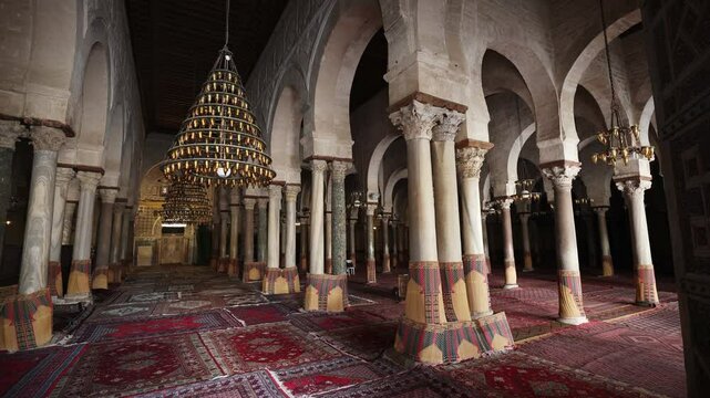 Forest of marble columns topped with carved Corinthian capitals in prayer hall of Kairouan Mosque of Uqba in Tunisia, covered with soft carpets and enlightened by chandeliers with small glass lamps