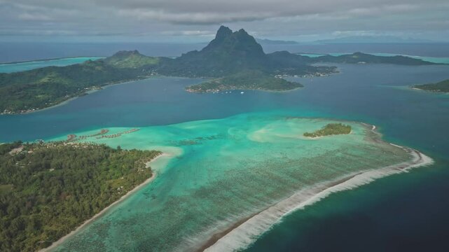 Aerial panorama vibrant coral reef in crystal clear, shallow water of Bora Bora turquoise lagoon, framed by majestic green mountain Otemanu rising dramatically in background. Exotic travel getaway.